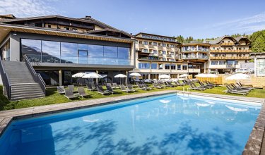 Hotel with pool and sunbathing area in front of forested mountains        Ask ChatGPT