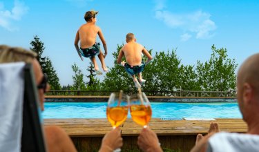 Two boys jump into the pool as two adults toast with drinks by the poolside