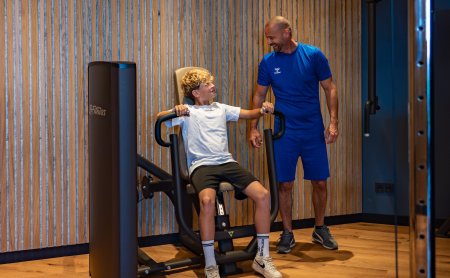 Young man exercises on fitness machine, trainer stands to the right smiling at him