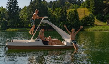 Familie auf einem Tretboot auf einem See, ein Junge klettert auf die Rutsche, ein anderer rutscht ins Wasser