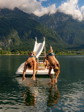 Zwei Jungen springen von einer Wasserrutsche auf einem See vor bewaldeten Bergen ins Wasser