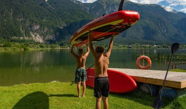 Zwei Jungen tragen ein rotes Stand-Up-Paddle-Board am Seeufer mit Bergen im Hintergrund