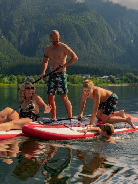 Family playing and laughing on and in the water with paddleboards in front of forested mountains