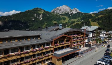 View of a hotel with flower-adorned balconies in front of forested mountains under blue sky