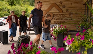 Familie mit Koffern betritt Hotel Garni Kofel, umgeben von Blumen und grüner Landschaft