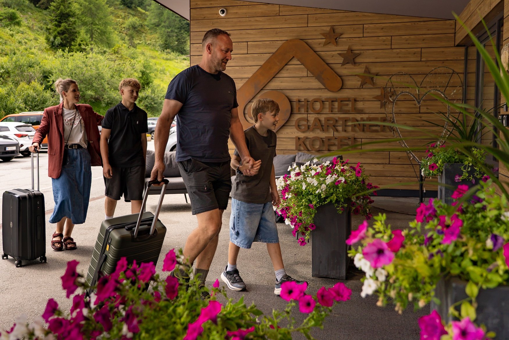 Family with suitcases entering Hotel Garni Kofel, surrounded by flowers and green landscape