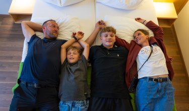 Familie liegt entspannt nebeneinander auf zwei Betten in einem Zimmer mit Holzboden