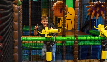 Junge spielt mit gelbem Spielgerät in einem bunten Indoor-Spielplatz hinter einem Netz