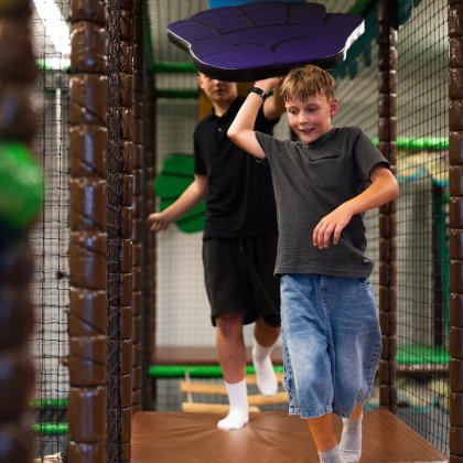 Junge läuft in einer Kletterhalle unter einem lila Hindernis, eine weitere Person im Hintergrund