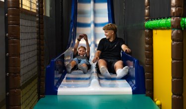 Zwei Jungen rutschen nebeneinander auf einer blauen Rutsche in einer Indoor-Spielhalle