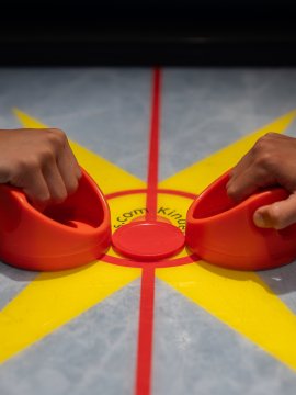 Hands gripping red paddles on an air hockey table with yellow star and red circle
