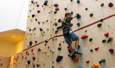 Kind klettert an einer bunten Kletterwand in einer Halle mit Sicherungsgurt