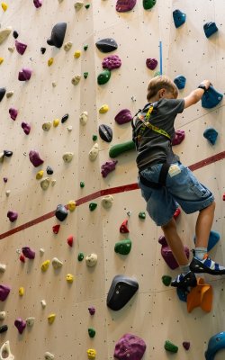 Kind klettert an einer bunten Kletterwand in einer Halle mit Sicherungsgurt