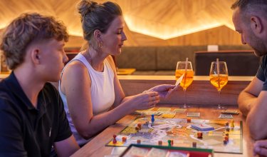 Drei Personen sitzen an einem Tisch und spielen ein Brettspiel mit Getränken in Gläsern
