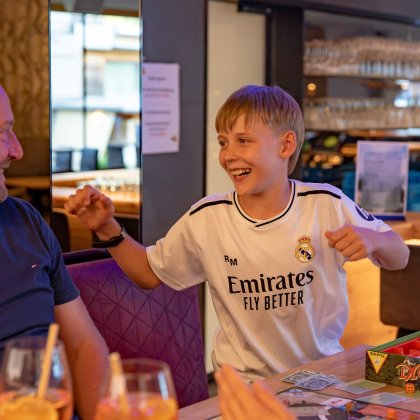 Junge in weißem Trikot lacht und gestikuliert gegenüber einem lächelnden Mann in einem Restaurant.