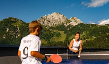 Zwei Personen spielen Tischtennis draußen mit Bergen und blauem Himmel im Hintergrund