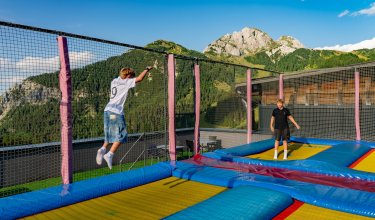 Zwei Jungen springen auf einem Trampolin mit Berglandschaft im Hintergrund unter blauem Himmel