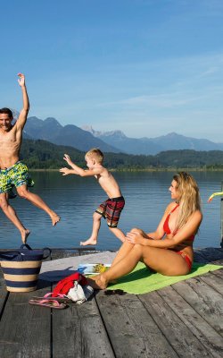 Familie am Badesteg, Kinder springen lachend in den See vor Bergkulisse