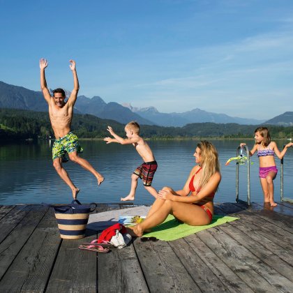 Familie am Badesteg, Kinder springen lachend in den See vor Bergkulisse