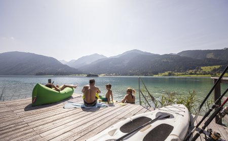 Familie am Steg mit Blick auf See und Berge, SUP und Angelruten im Vordergrund