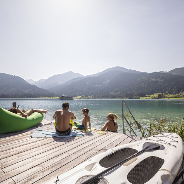 Familie am Steg mit Blick auf See und Berge, SUP und Angelruten im Vordergrund