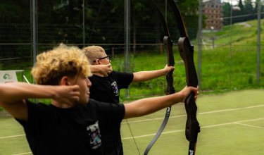 Zwei Jungen stehen nebeneinander auf einem Sportplatz und zielen mit Bögen nach rechts.