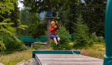Person wearing helmet and harness riding a zip line over a wooden platform in the forest