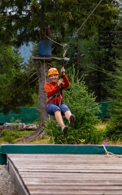 Person mit Helm und Kletterausrüstung fährt auf einer Seilrutsche über eine Holzplattform im Wald