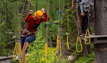 Person klettert an Seilbrücke mit gelben Schlaufen, andere warten auf Plattform im Wald