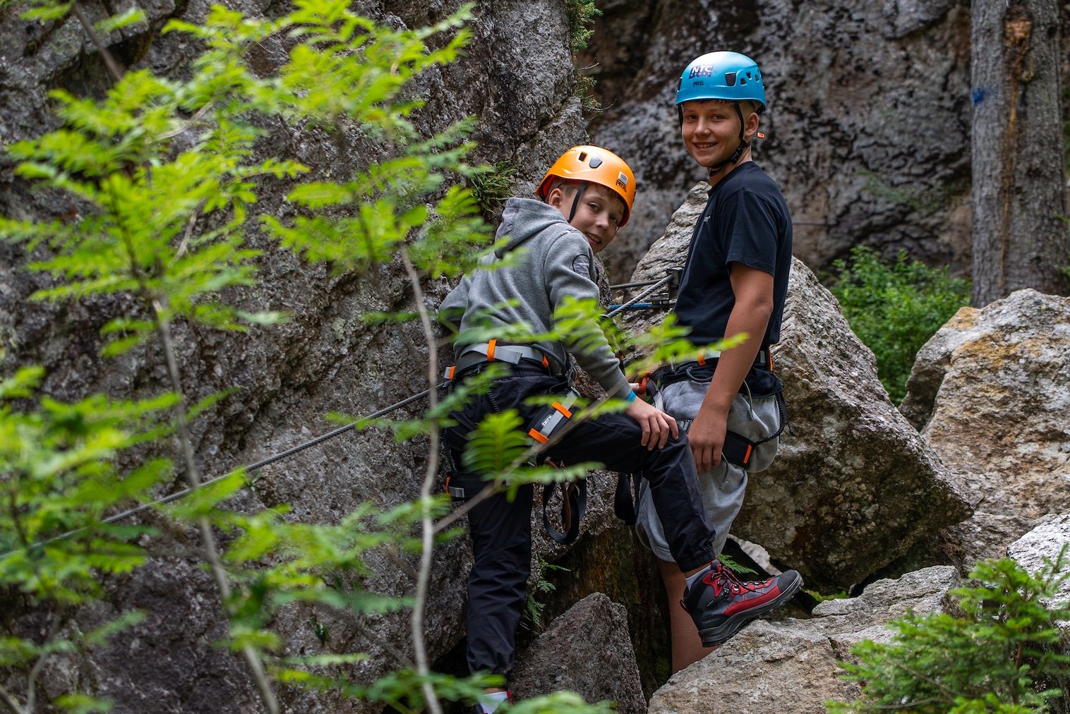 Two teens wearing climbing gear and helmets standing on rocks in a forest