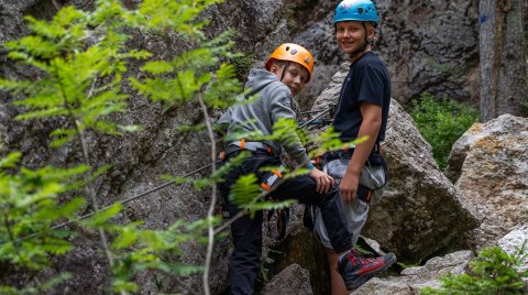 Two teens wearing climbing gear and helmets standing on rocks in a forest