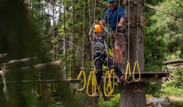 Kind mit Helm klettert an Seilparcours, Erwachsener sichert von Plattform im Wald