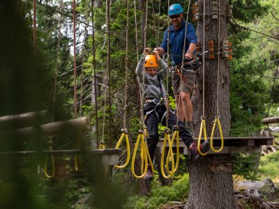 Kind mit Helm klettert an Seilparcours, Erwachsener sichert von Plattform im Wald