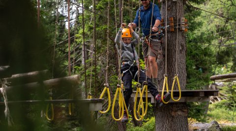 Child wearing helmet climbs rope course while adult secures from platform in forest