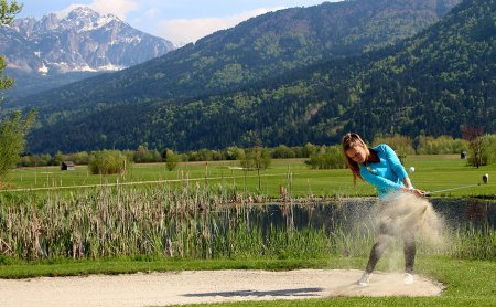 Golferin schlägt Ball aus dem Bunker vor Bergkulisse