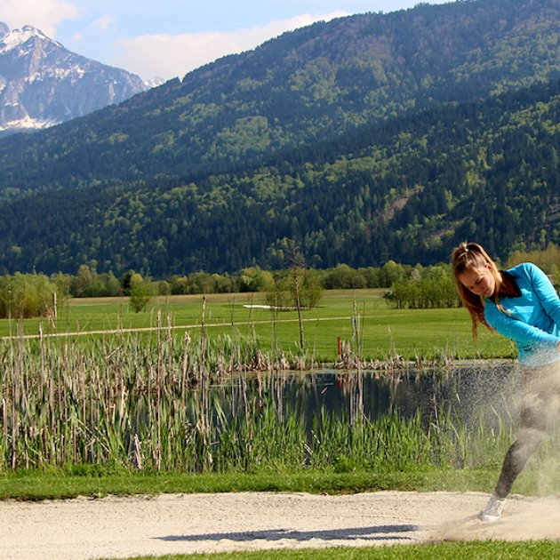 Golferin schlägt Ball aus dem Bunker vor Bergkulisse