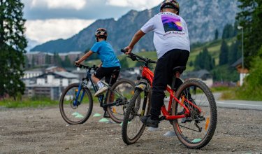 Zwei Kinder mit Helmen fahren Mountainbikes auf einem Schotterweg vor Bergkulisse.