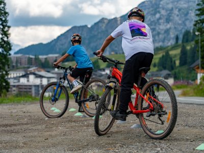 Zwei Kinder mit Helmen fahren Mountainbikes auf einem Schotterweg vor Bergkulisse.