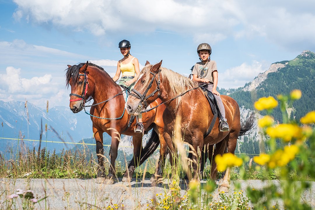 Zwei Kinder reiten auf braunen Pferden in alpiner Landschaft