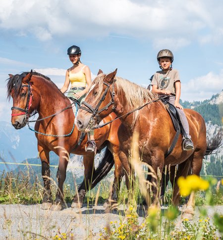 Zwei Kinder reiten auf braunen Pferden in alpiner Landschaft