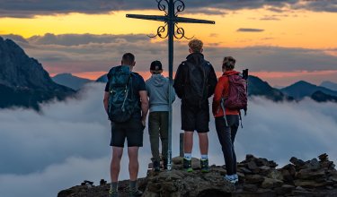 Vier Wanderer stehen vor einem Gipfelkreuz bei Sonnenuntergang über den Wolken in den Bergen
