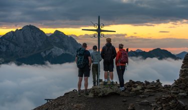 Four hikers stand by a summit cross looking at mountains and clouds at sunset