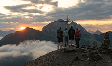 Four hikers stand at a summit cross at sunset with mountain views and clouds