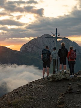 Four hikers stand at a summit cross at sunset with mountain views and clouds