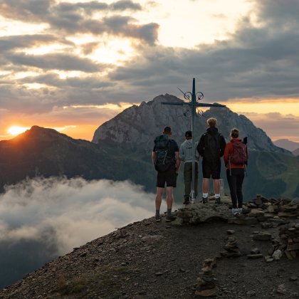Vier Wanderer stehen bei Sonnenuntergang an einem Gipfelkreuz mit Bergpanorama und Wolken