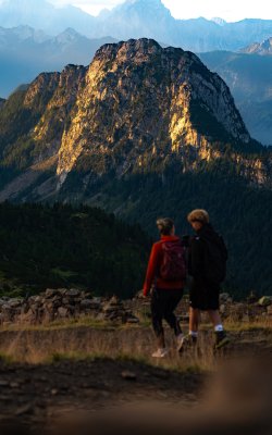 Two hikers in the foreground looking at a sunlit mountain with forested slopes