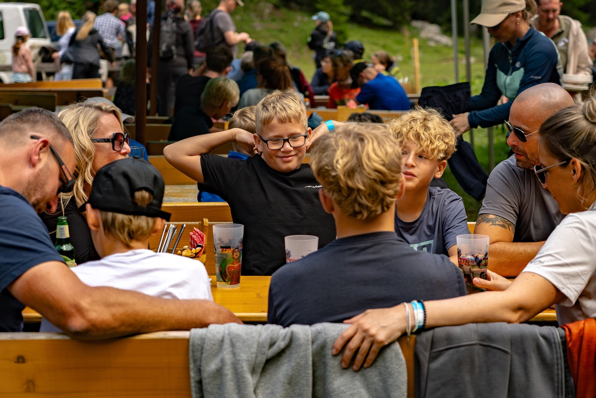 Gruppe von Menschen sitzt an Holztischen im Freien, Kinder und Erwachsene unterhalten sich