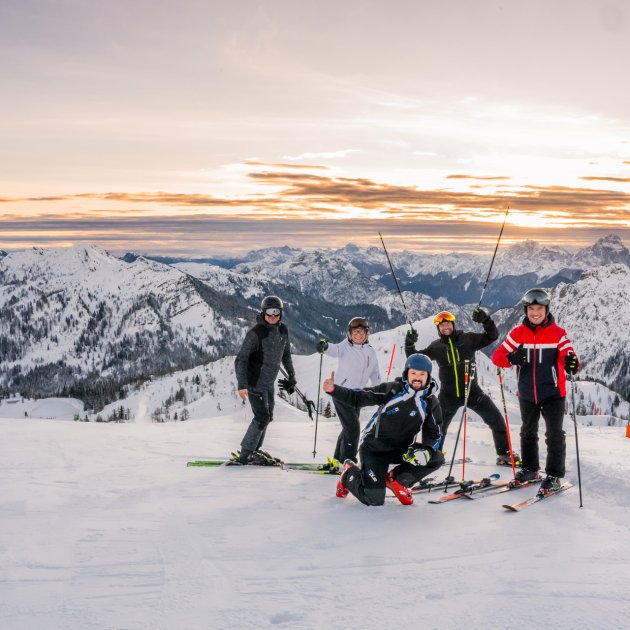 Fünf Skifahrer posieren zusammen auf der Piste bei Sonnenaufgang, Berge im Hintergrund.