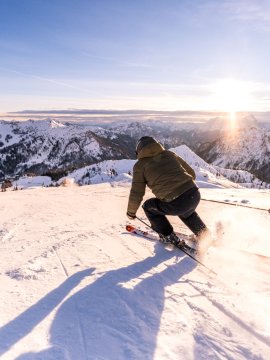 Ein Skifahrer fährt die Piste hinab, die Sonne geht hinter den Bergen auf.