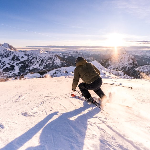 Ein Skifahrer fährt die Piste hinab, die Sonne geht hinter den Bergen auf.